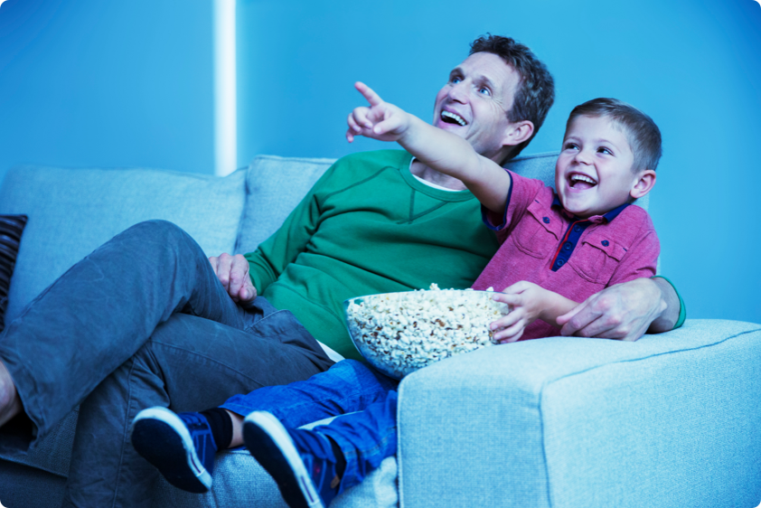 Father and son smiling and watching television on sofa with bowl of popcorn.